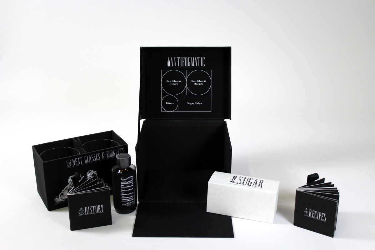 An opened black box displayed with all contents within: 2 neat glasses, a box of sugar cubes, a bottle of angostura bitters, and two small recipe books