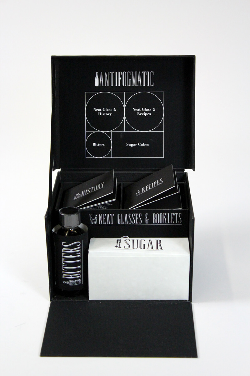 A black box with a box of sugar, two booklets, and a bottle of bitters.